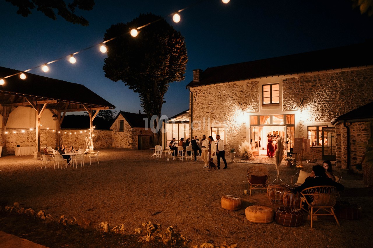 Cour d'une maison en pierre éclairée par des guirlandes lumineuses, avec des invités assis et debout en soirée.