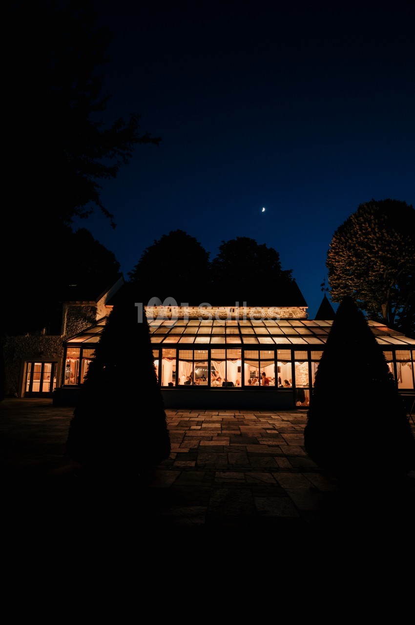 Verrière lumineuse d'une maison entourée d'arbres, éclairée de nuit sous un ciel étoilé avec une lune visible.