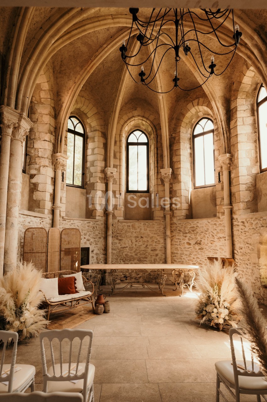 Salle voûtée en pierre avec grandes fenêtres, décorée de fleurs séchées, chaises blanches et table en bois.