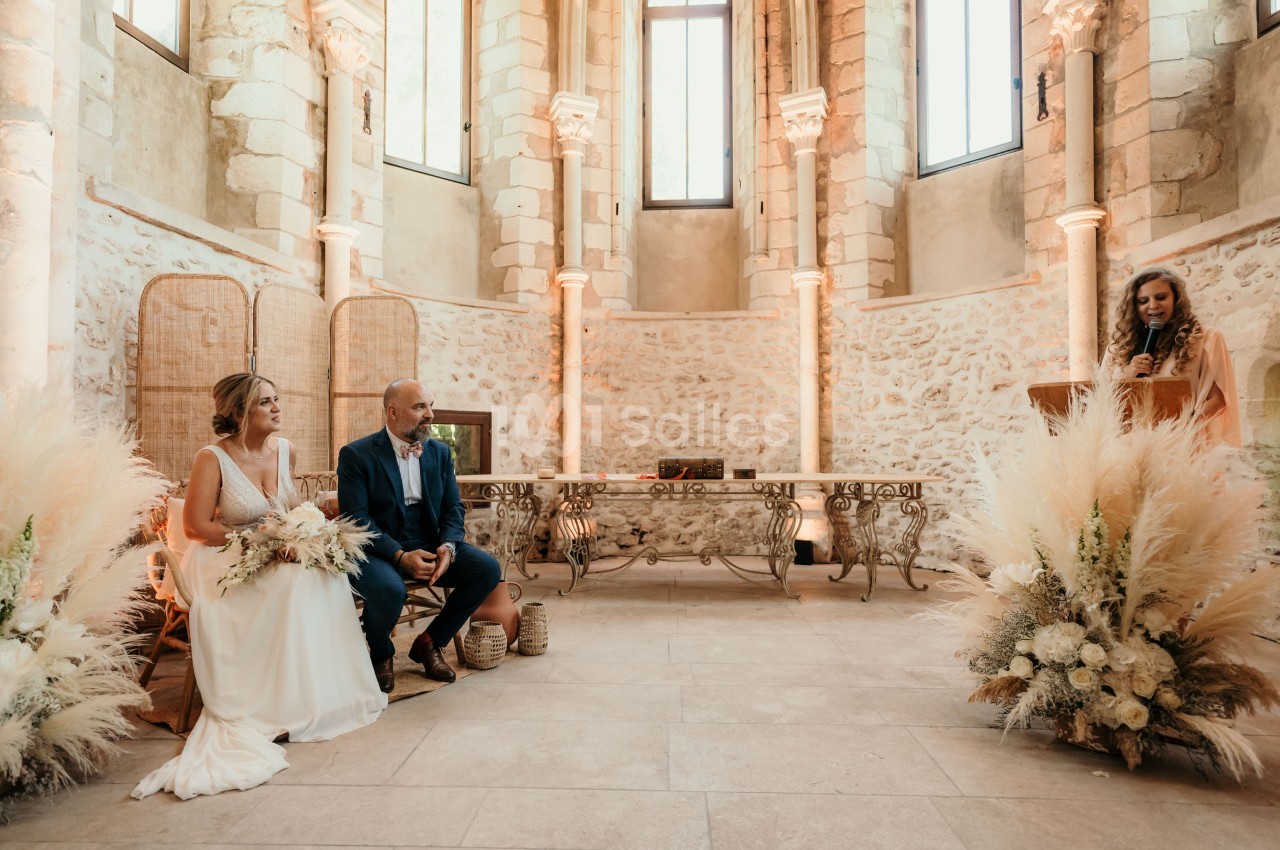 Un couple assis dans une salle lumineuse au style ancien, écoutant une femme debout à un pupitre.