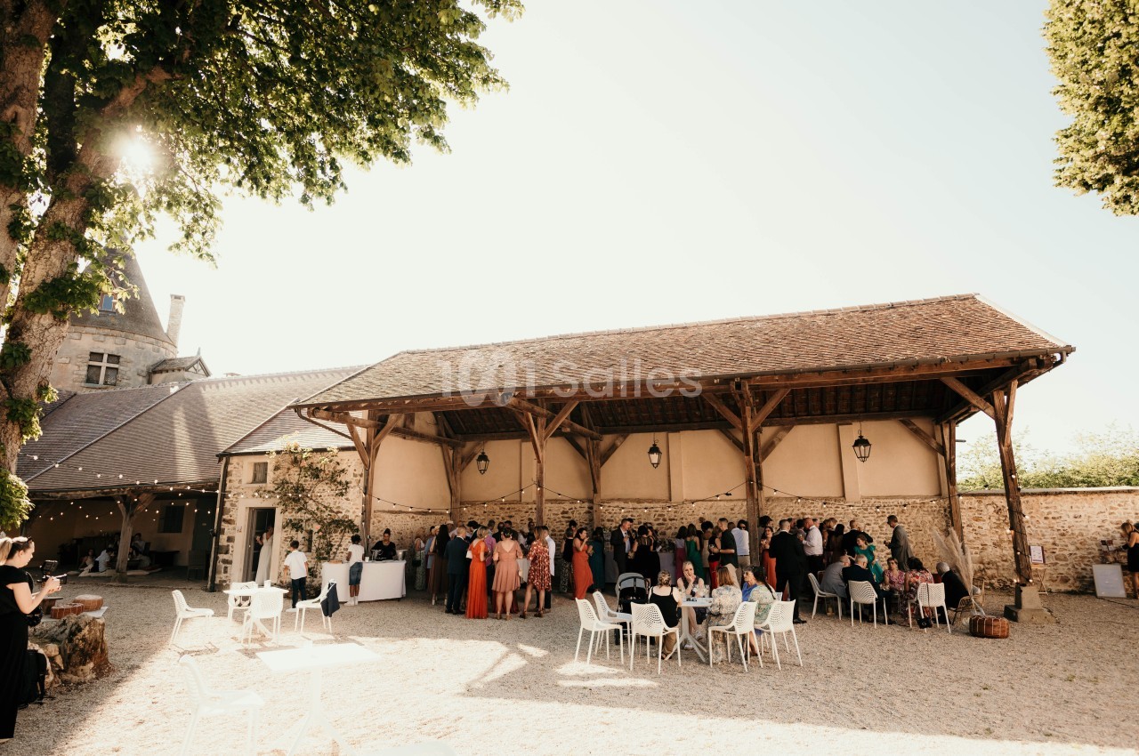 Groupe de personnes rassemblées sous une grande structure en bois dans une cour ensoleillée, avec des tables et des chaises.