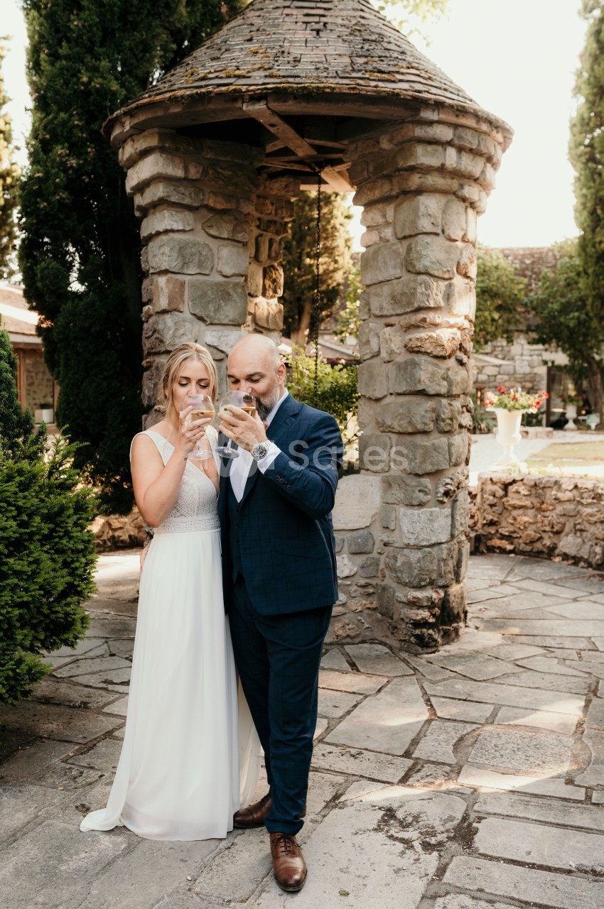 Un couple en tenue de mariage boit un verre devant un puits en pierre dans un jardin.