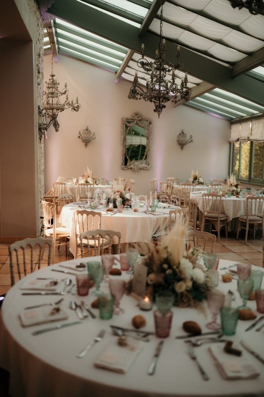 Salle de réception lumineuse avec tables décorées de fleurs, vaisselle colorée et chandeliers suspendus.