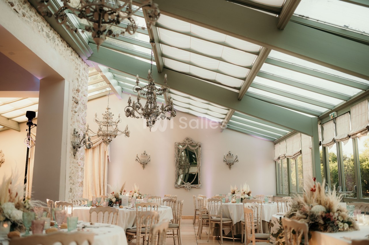 Salle de réception lumineuse avec verrière, tables dressées et décorations élégantes dans des tons clairs.
