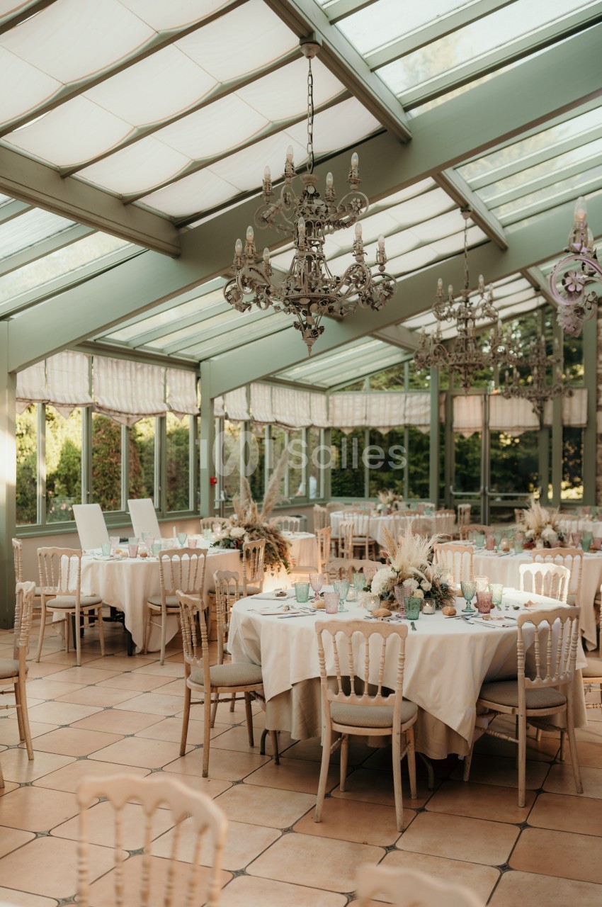 Salle lumineuse avec verrière, tables rondes dressées pour un événement, décorations florales et lustres suspendus.