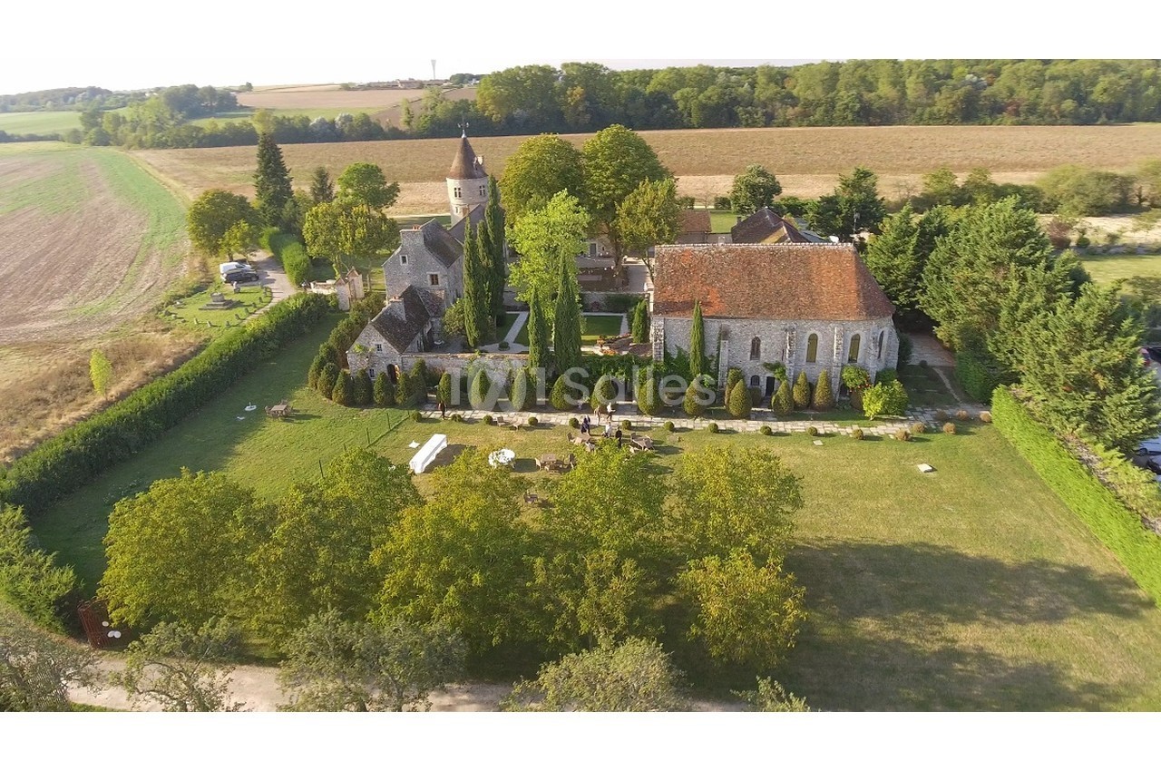 Vue aérienne d'un domaine rural avec une église, des bâtiments en pierre, un jardin arboré et des champs environnants.