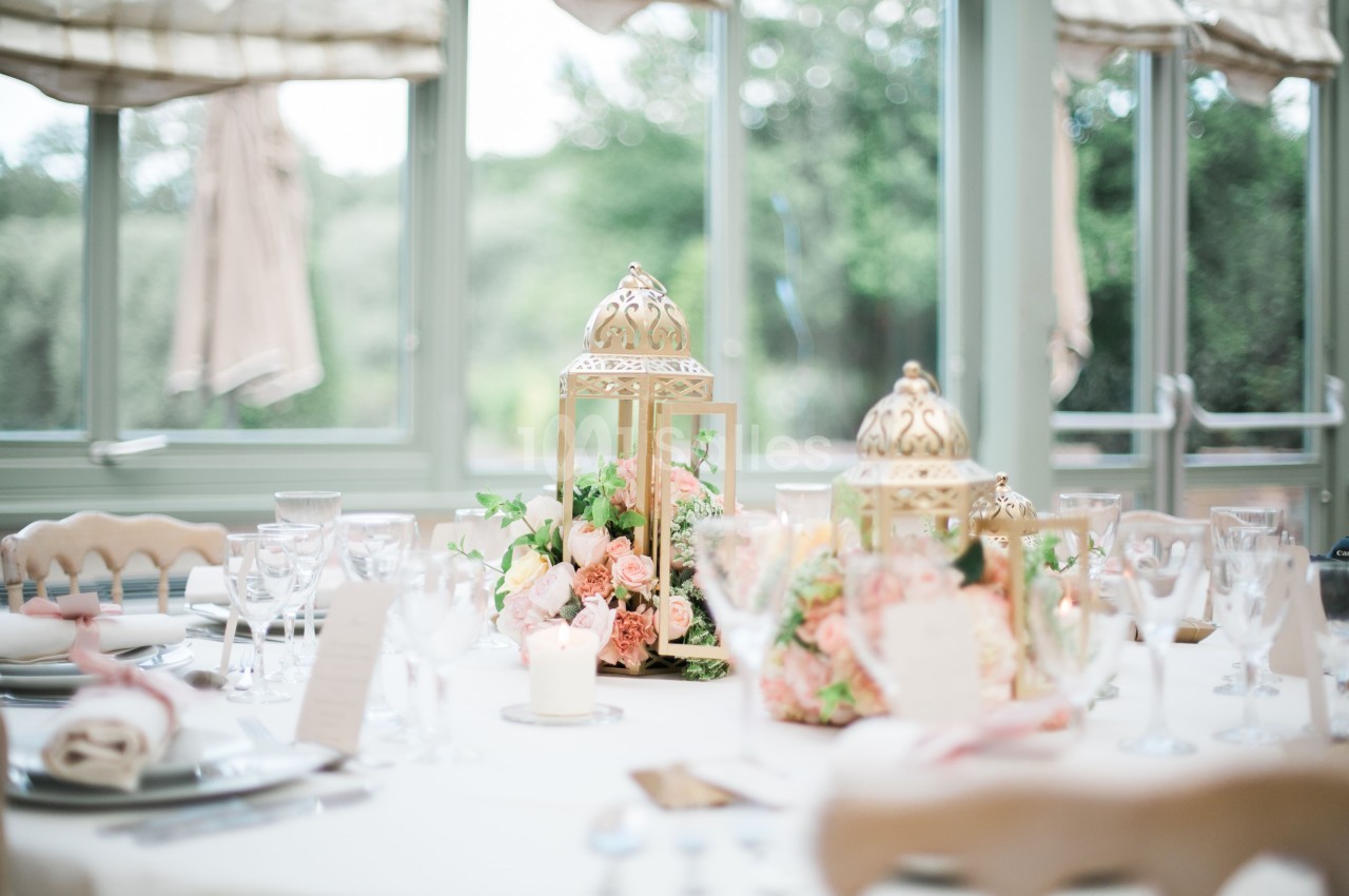 Table décorée avec des lanternes dorées, des fleurs roses et des verres, dans une salle lumineuse avec vue sur un jardin.