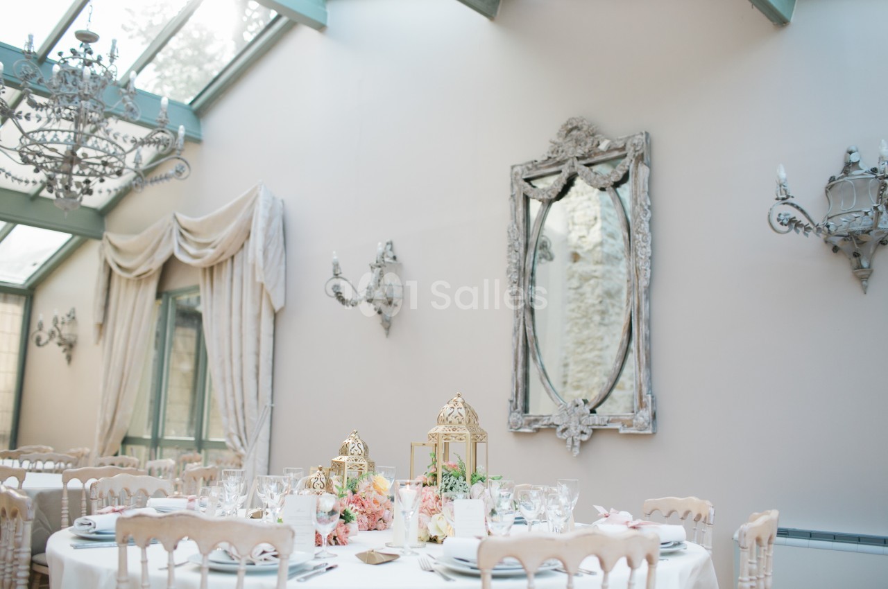 Salle lumineuse avec table décorée de fleurs et lanternes dorées, entourée de chaises et ornée de miroirs et lustres.