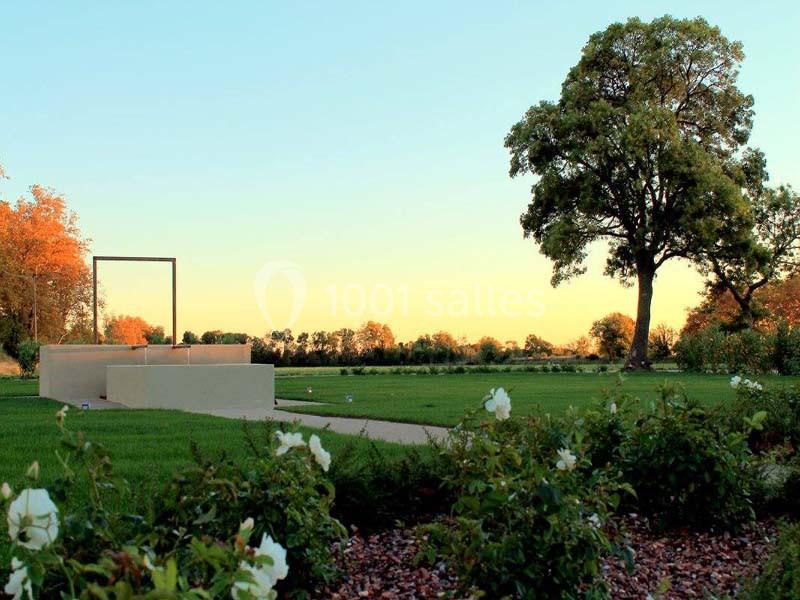 Jardin paysager avec pelouse, rosiers, arbre imposant et structure moderne, sous un ciel dégagé au coucher du soleil.