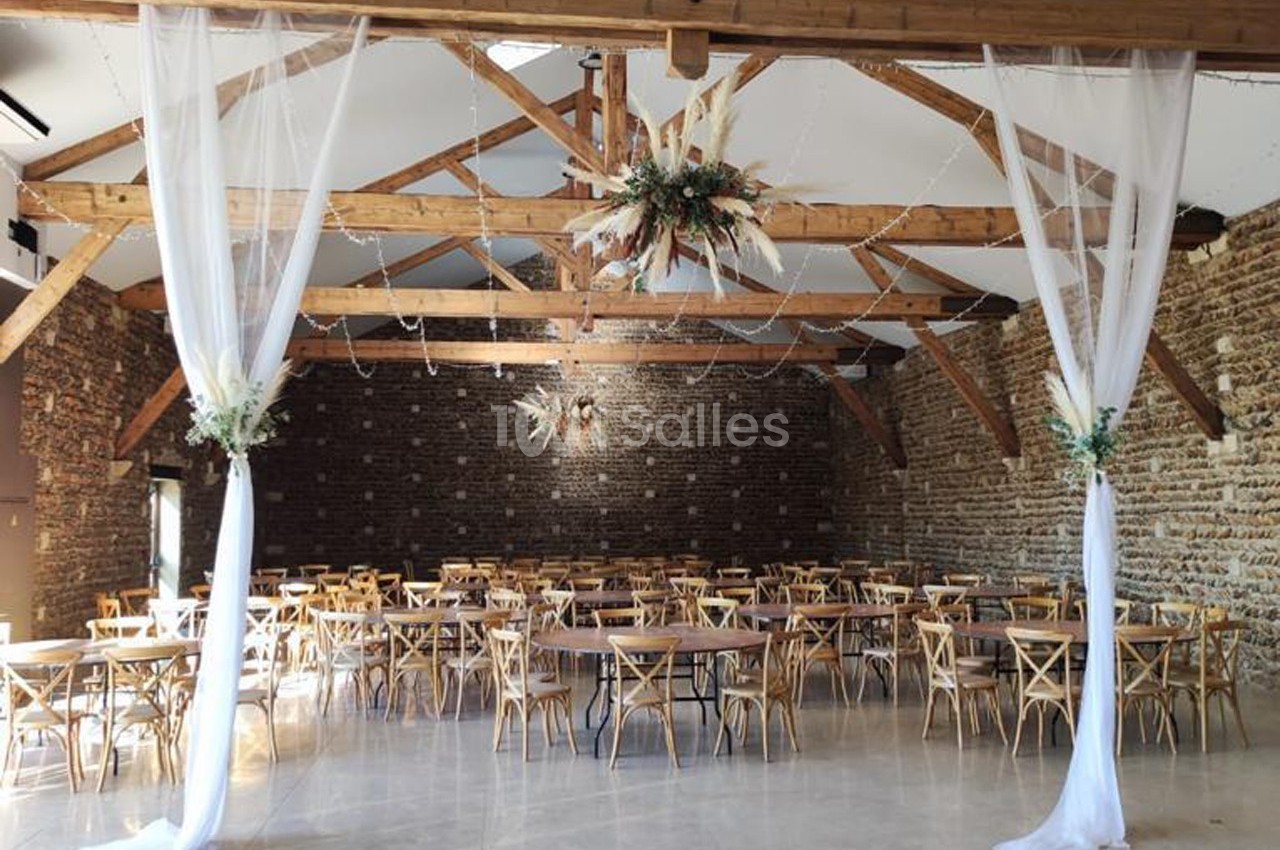 Salle de réception avec murs en briques, poutres apparentes, tables en bois et décorations florales suspendues.
