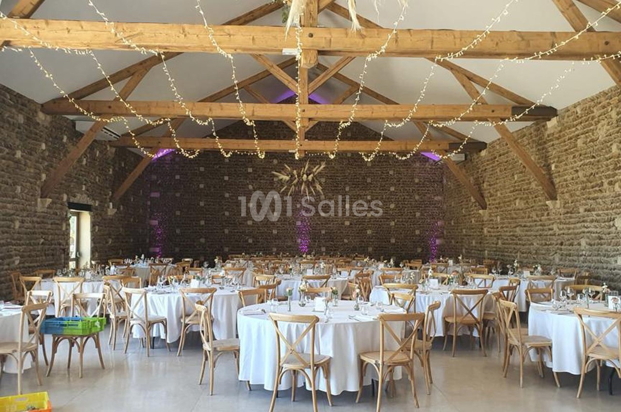 Salle de réception en pierre avec tables rondes dressées, chaises en bois et guirlandes lumineuses suspendues.