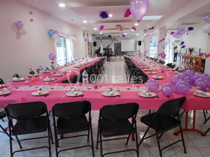 Salle décorée pour une fête avec des tables alignées, nappes roses, ballons violets et assiettes dressées.