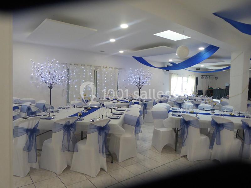 Salle de réception décorée avec des tables dressées, des chaises blanches ornées de nœuds bleus et des arbres lumineux.