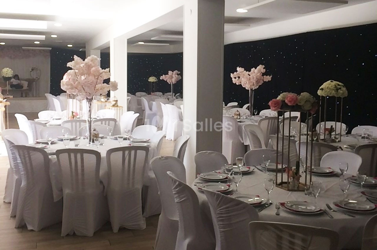 Salle de réception décorée avec des tables rondes, nappes blanches, chaises assorties et centres de table fleuris.