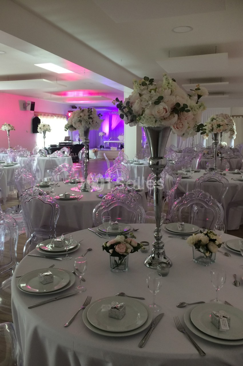 Salle de réception décorée avec des tables rondes, nappes blanches, centres de table floraux et chaises transparentes.