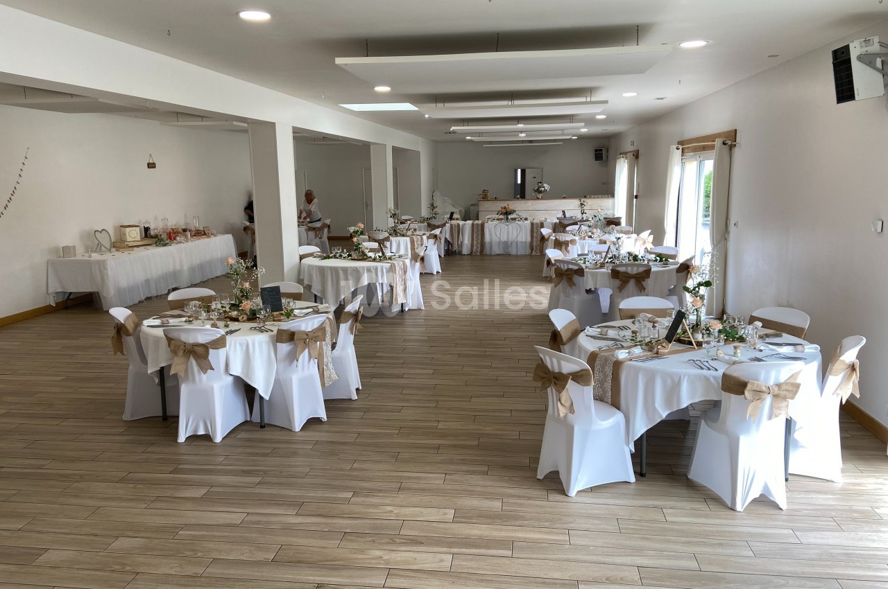 Salle de réception décorée pour un événement, avec des tables rondes dressées et des chaises ornées de nœuds beiges.
