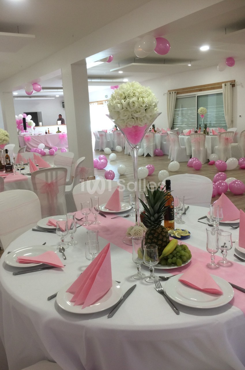 Salle décorée pour un événement avec tables dressées, nappes blanches, serviettes roses et ballons roses et blancs.