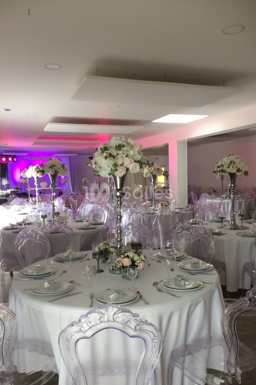 Salle de réception décorée avec des tables rondes, nappes blanches, chaises transparentes et centres de table fleuris.