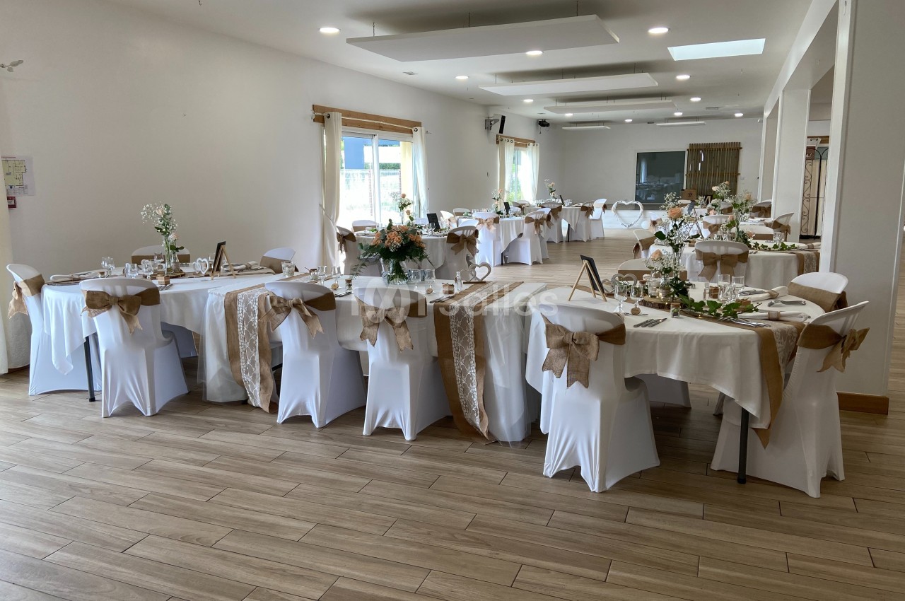 Salle de réception décorée pour un événement, avec tables dressées, nappes blanches et ornements beige et verts.