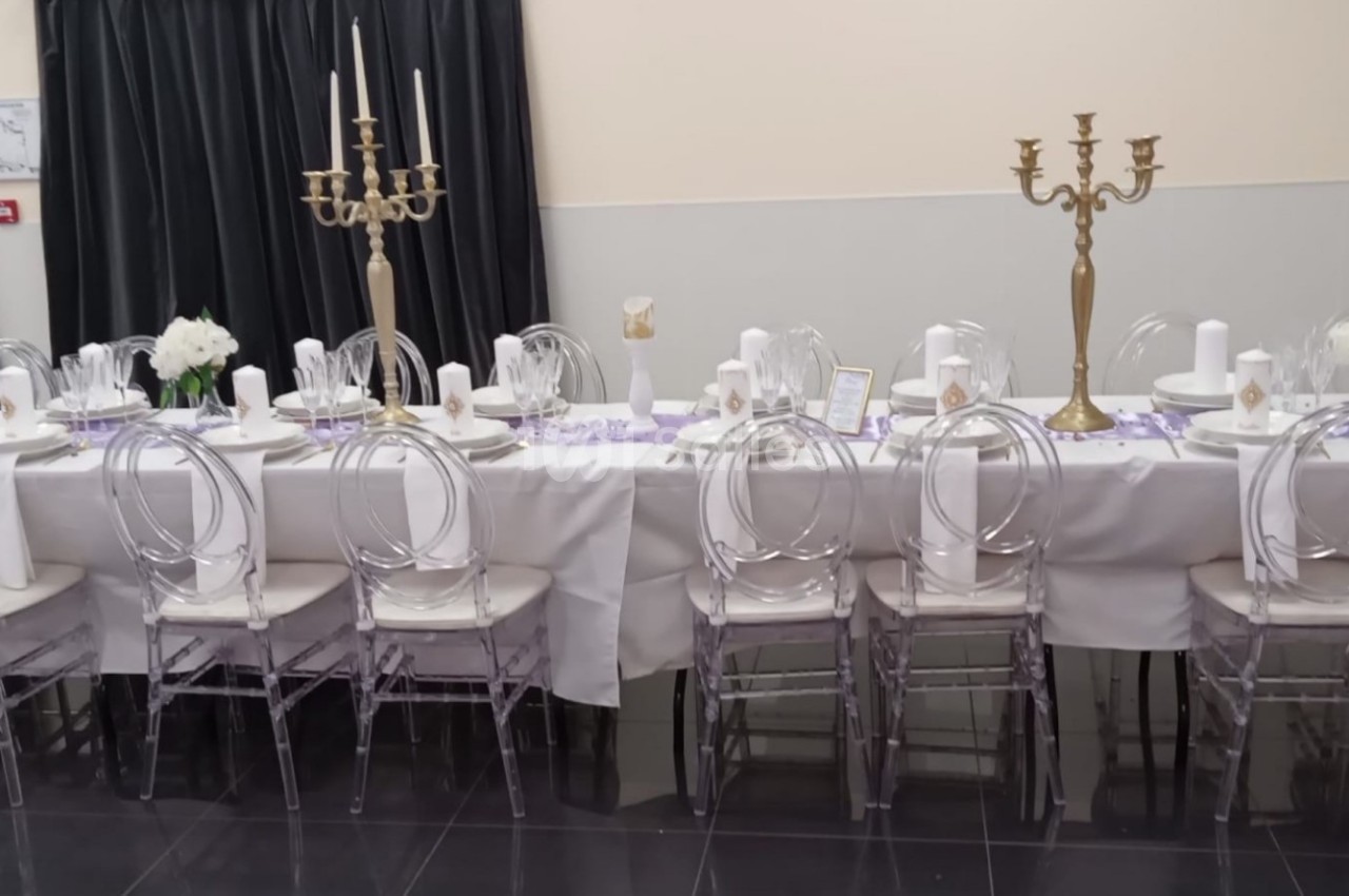 Table dressée pour un événement avec nappes blanches, chaises transparentes, chandeliers dorés et décorations florales.