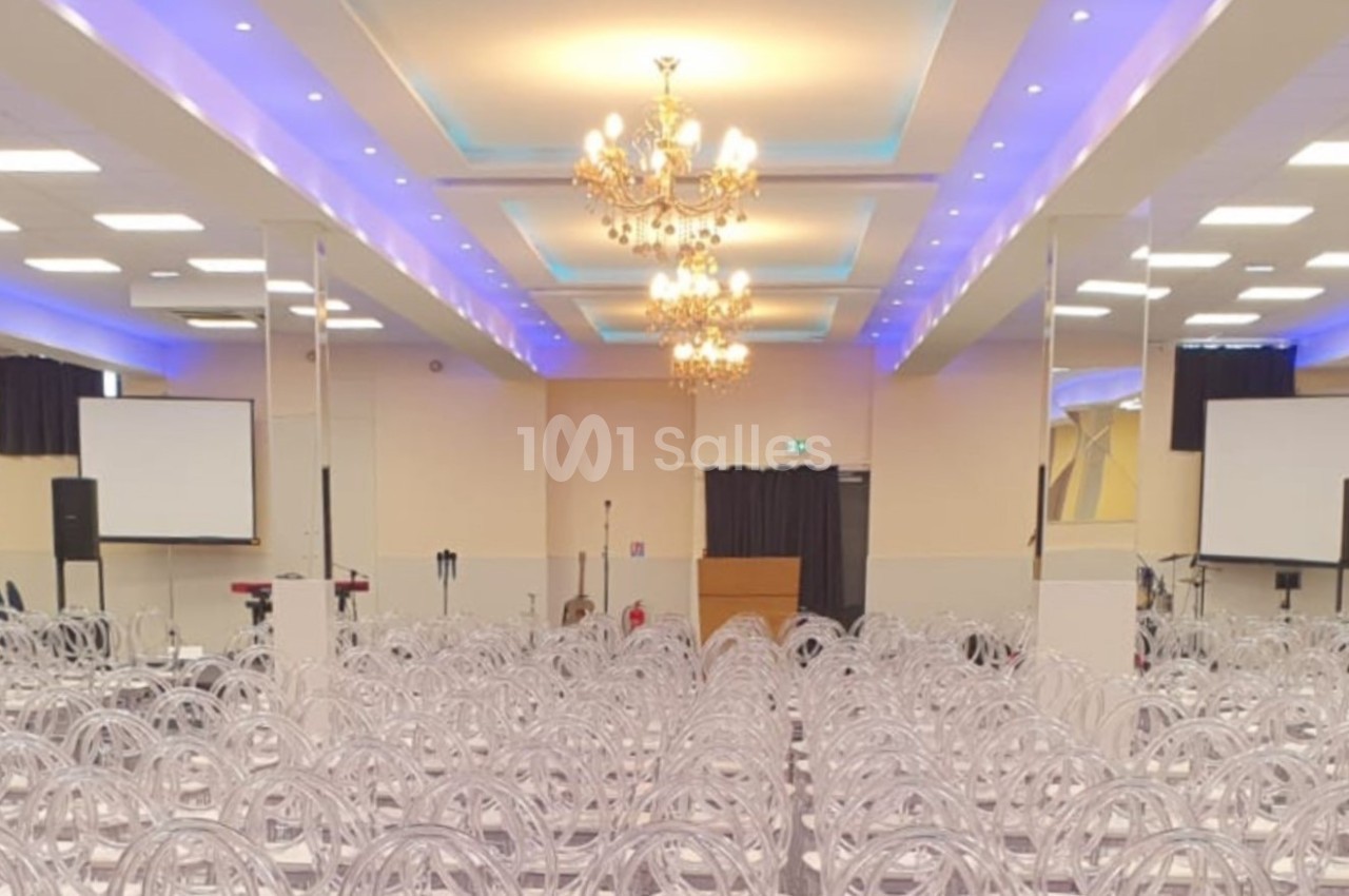 Salle de conférence lumineuse avec rangées de chaises transparentes, écrans de projection et lustres au plafond.