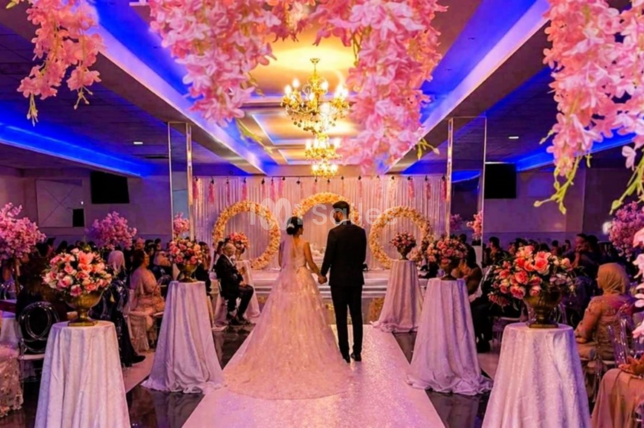 Un couple marche dans une allée décorée de fleurs roses sous un éclairage coloré lors d'une cérémonie de mariage.