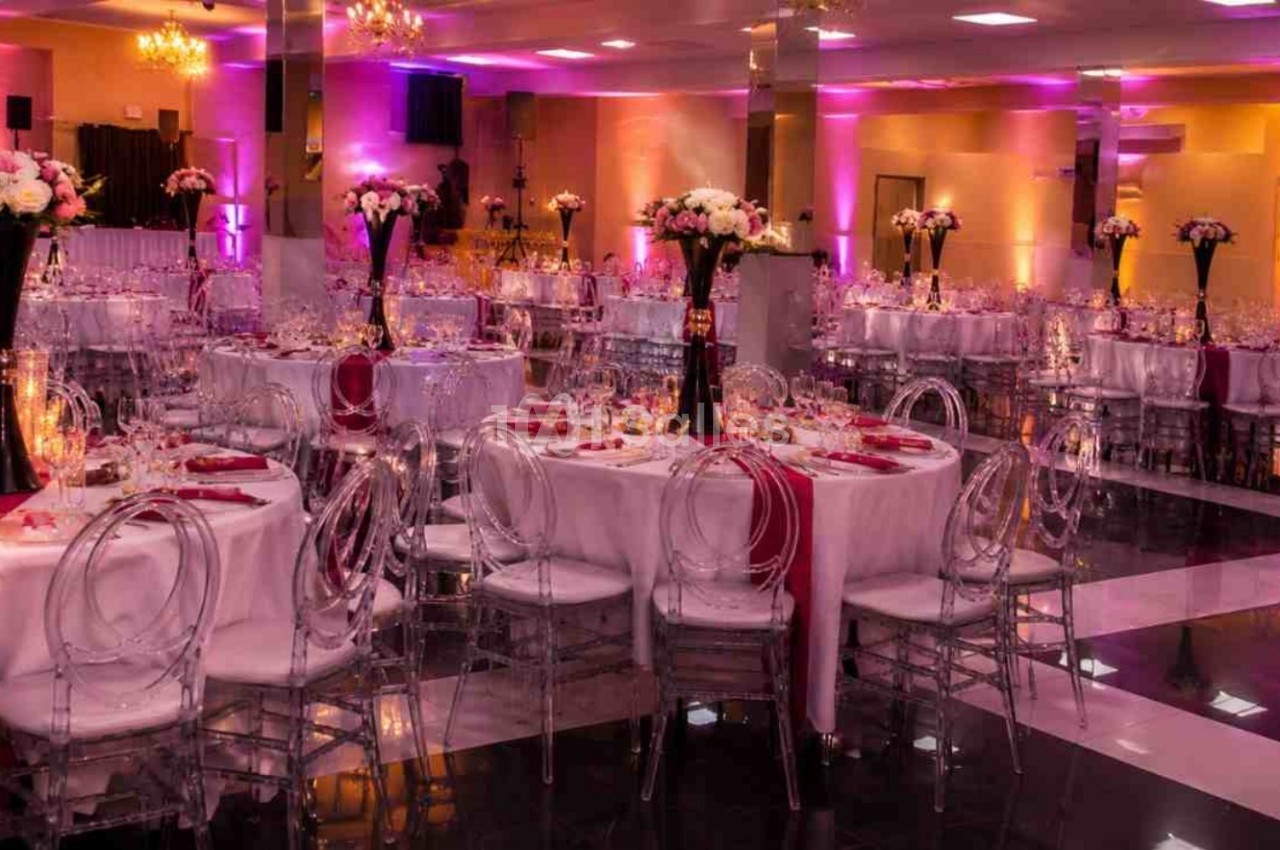 Salle de réception décorée avec des tables rondes, nappes blanches, chaises transparentes et éclairage rose.