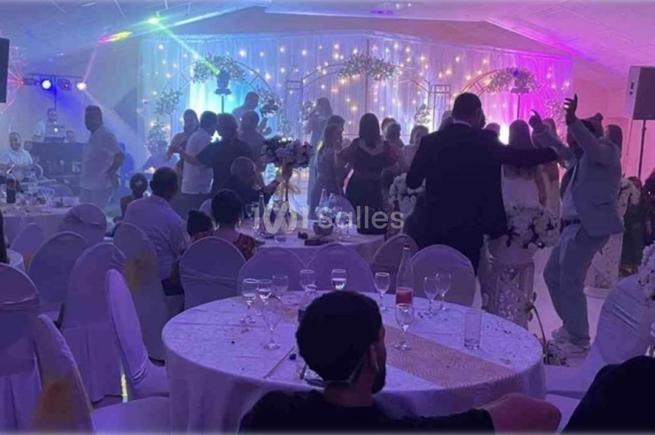 Salle de réception décorée, invités dansant et discutant autour de tables dressées, éclairage festif en arrière-plan.