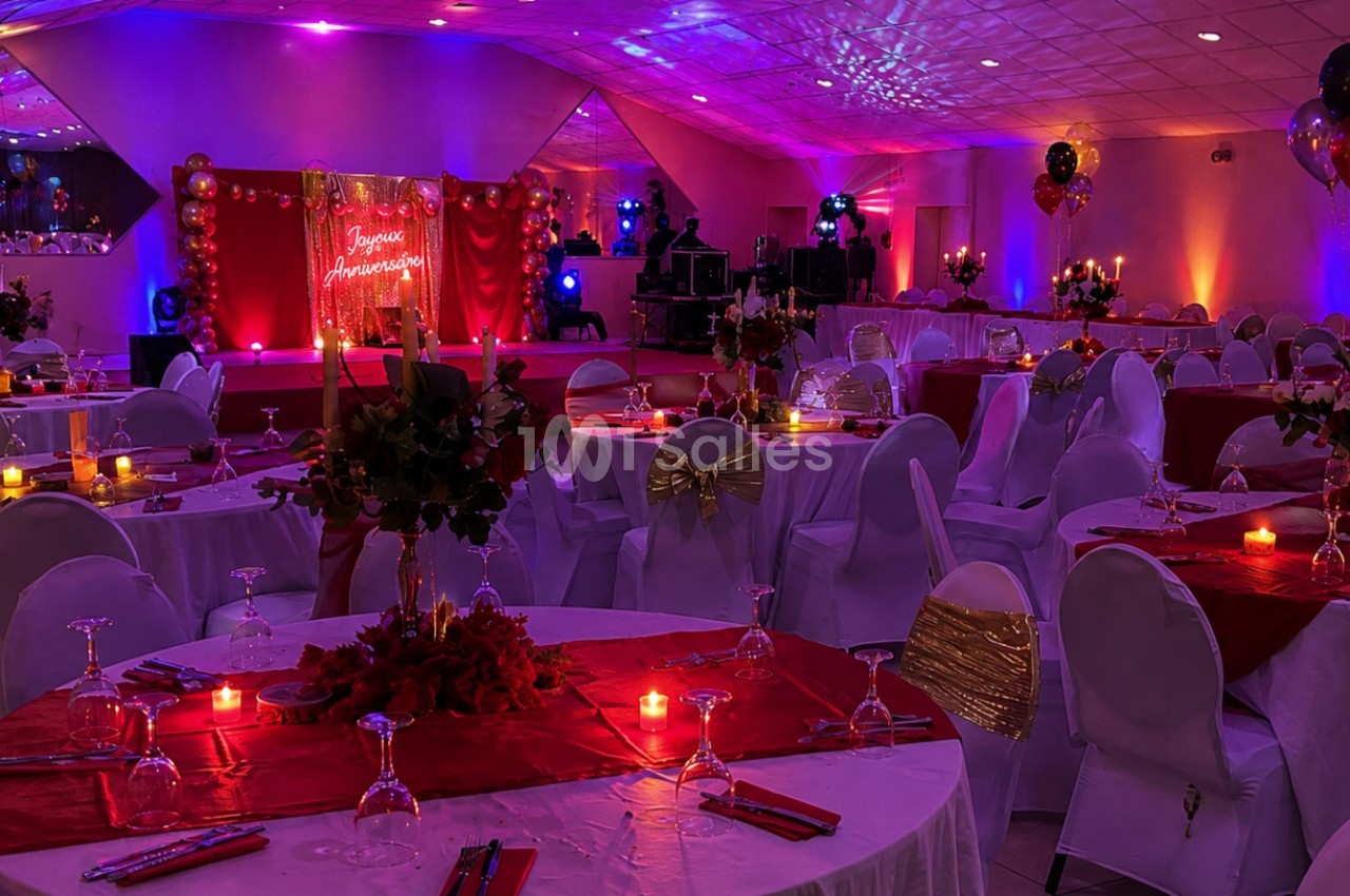 Salle de réception décorée avec des nappes rouges, bougies, ballons et éclairage tamisé pour un événement festif.
