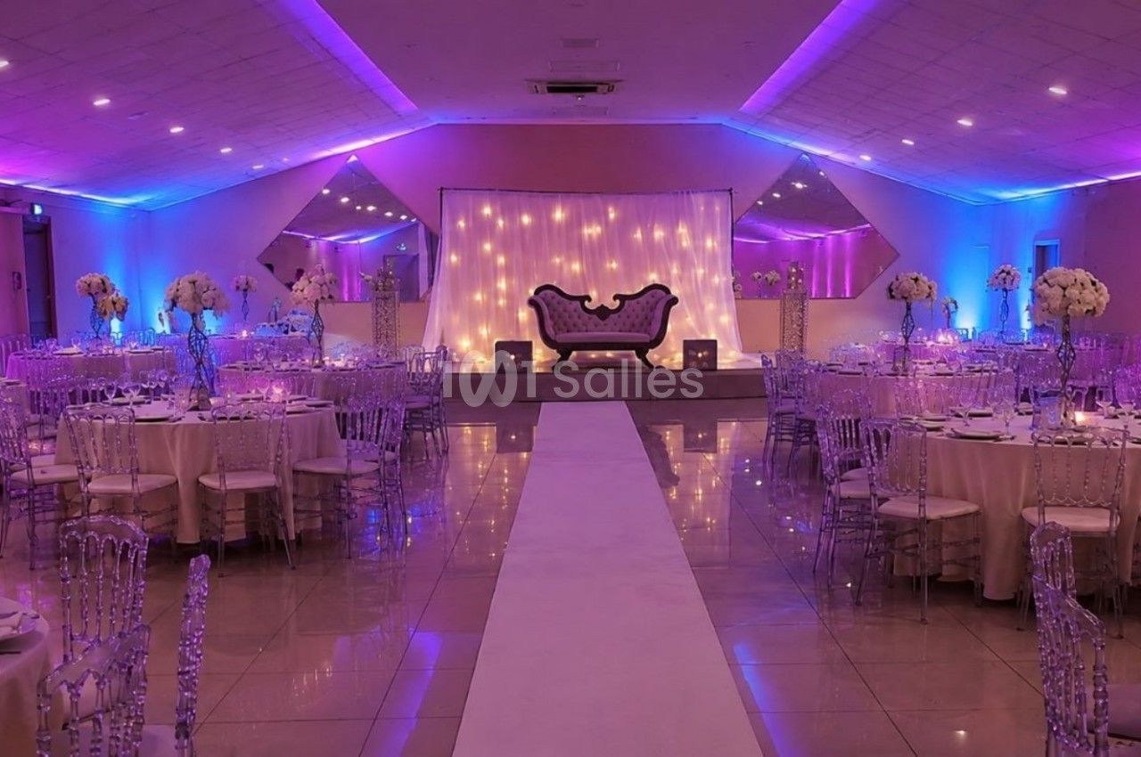 Salle de réception décorée avec des tables rondes, des chaises transparentes et un éclairage violet et bleu.