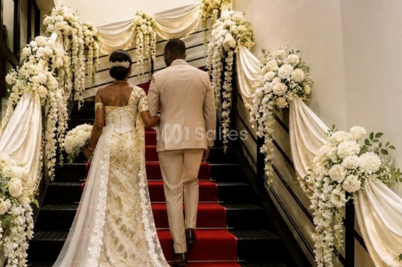 Un couple en tenue de mariage monte un escalier décoré de fleurs blanches et de drapés élégants.