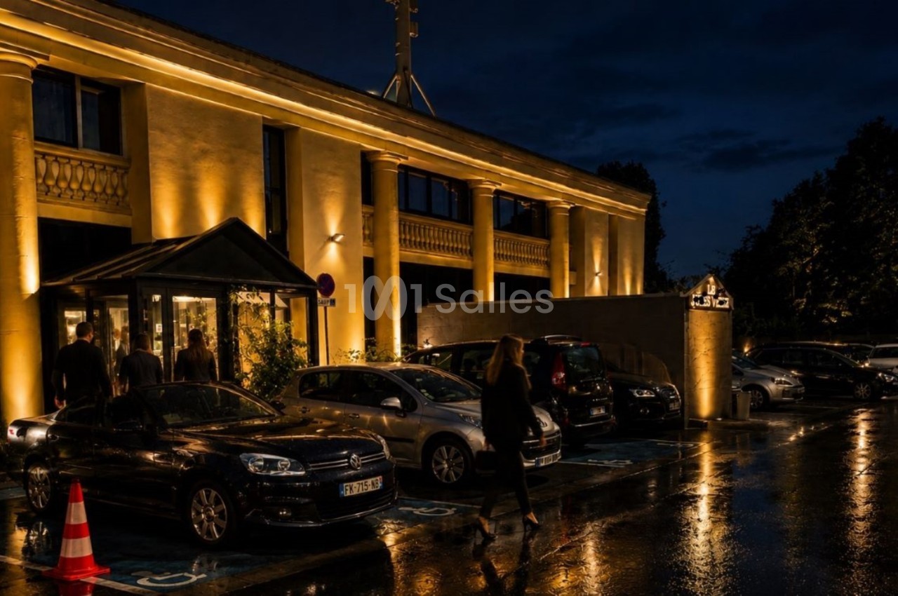 Façade d'un bâtiment éclairé de nuit avec des voitures garées sur un parking mouillé par la pluie.