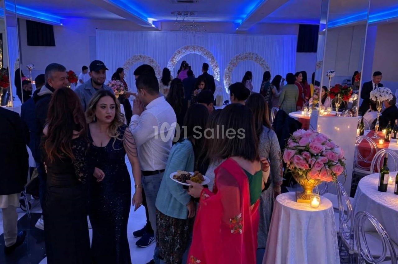 Salle de réception décorée avec des lumières bleues, des invités discutant autour de tables ornées de fleurs et bougies.