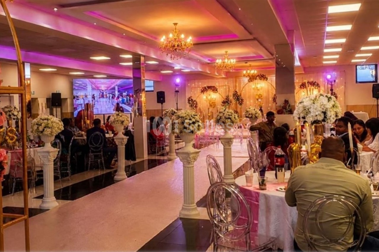Salle de réception décorée avec des fleurs et éclairages colorés, des invités assis à des tables rondes.