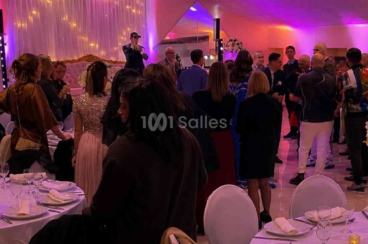 Des invités debout dans une salle de réception décorée avec des fleurs et des lumières colorées, écoutant un discours.