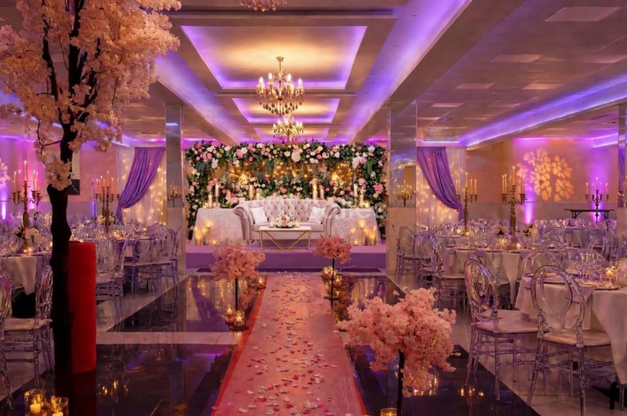 Salle de réception décorée avec des fleurs, des chandeliers et un éclairage violet, avec une allée centrale ornée de pétales.