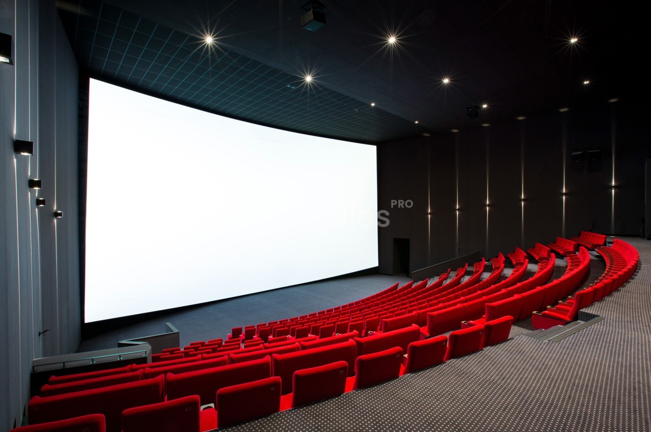 Salle de cinéma moderne avec des rangées de sièges rouges et un grand écran blanc allumé.