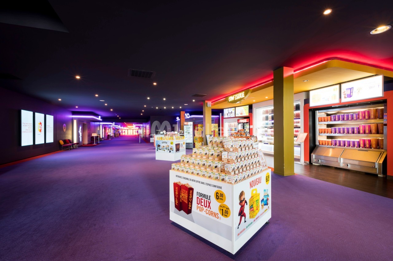 Présentoirs de confiseries et boissons dans le hall lumineux d'un cinéma avec éclairage coloré.