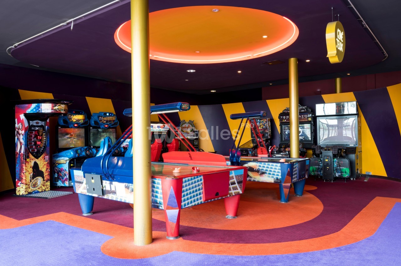 Salle d'arcade colorée avec des machines de jeux, des tables de hockey sur air et un éclairage néon au plafond.