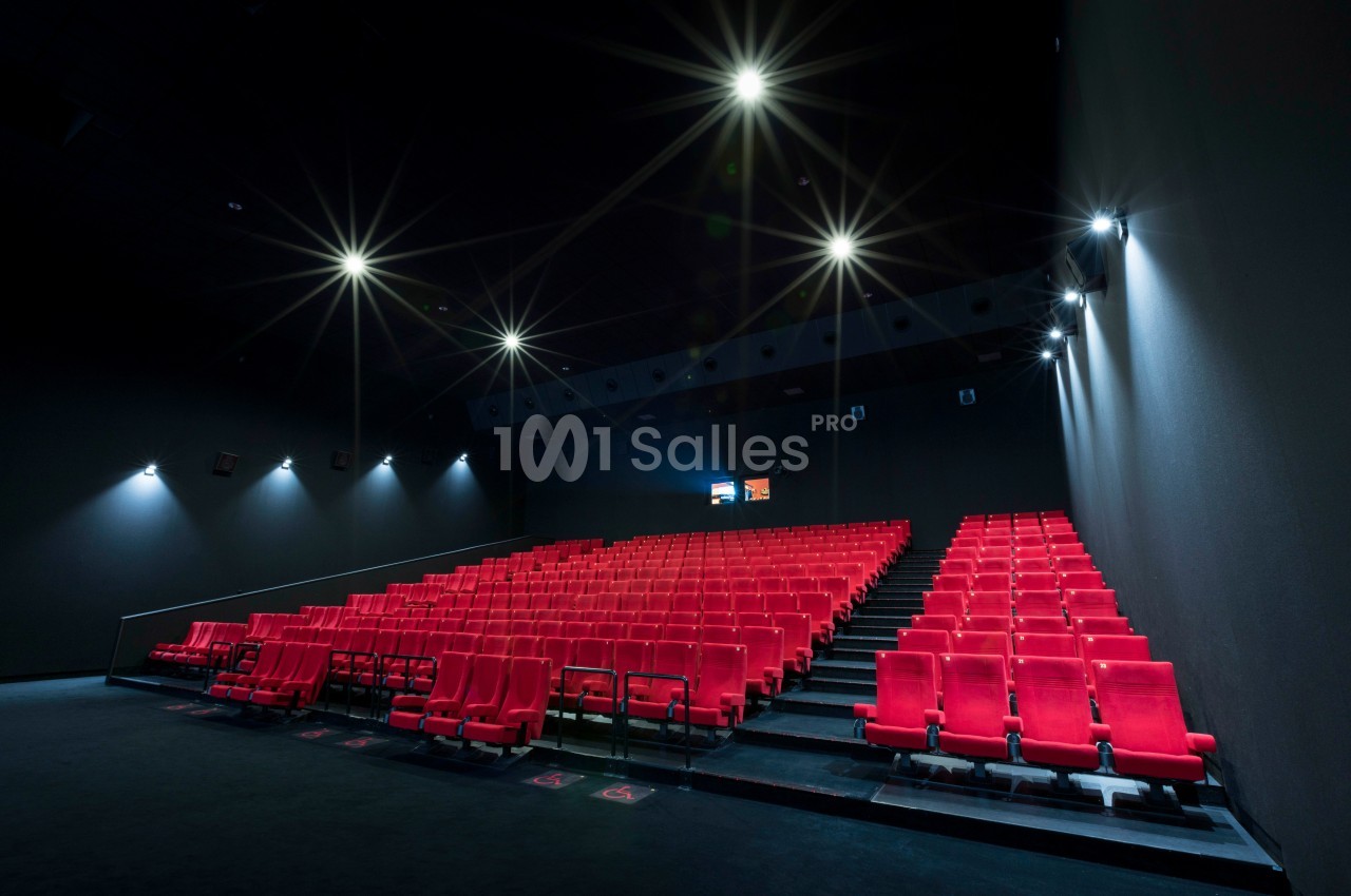 Salle de cinéma vide avec des rangées de sièges rouges et un écran allumé au fond.