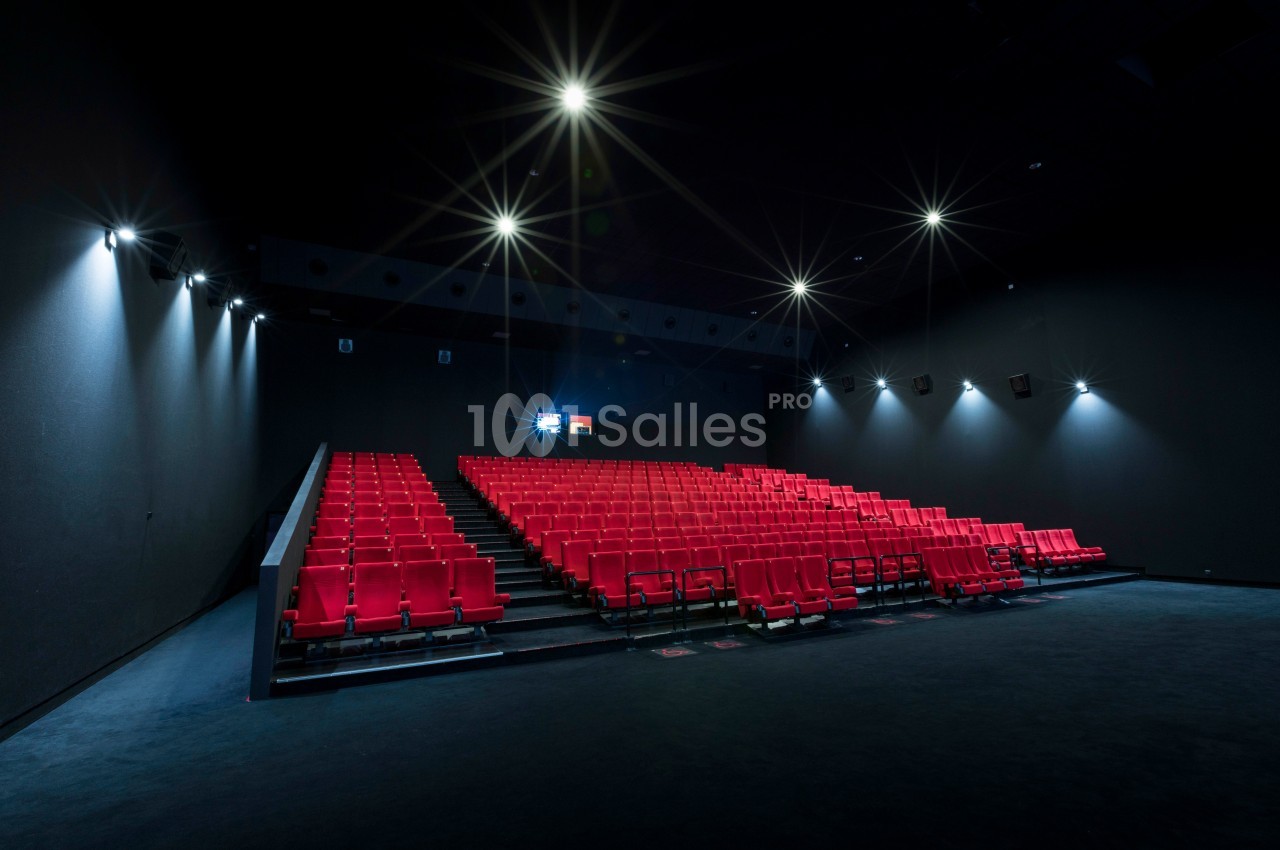 Salle de cinéma vide avec des rangées de sièges rouges et un écran allumé au fond.