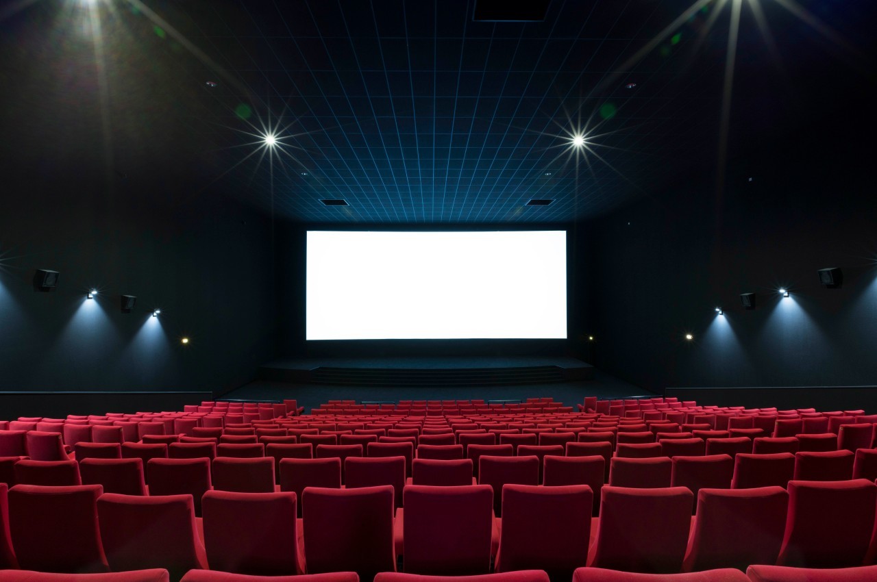 Salle de cinéma vide avec des rangées de sièges rouges et un écran blanc allumé au fond.