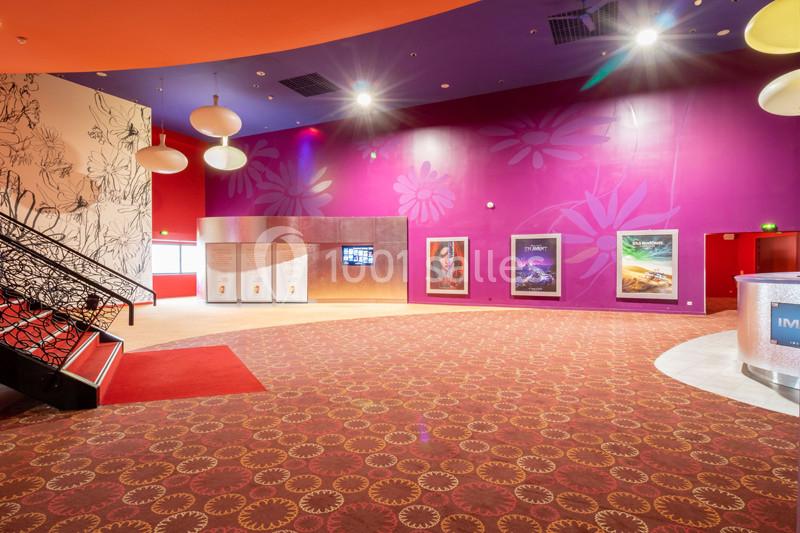 Hall de cinéma coloré avec moquette à motifs, affiches de films sur un mur violet et escalier décoratif à gauche.