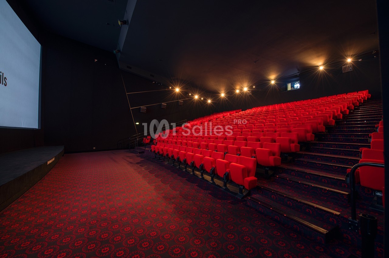Salle de cinéma vide avec des rangées de sièges rouges et un écran blanc allumé à gauche.
