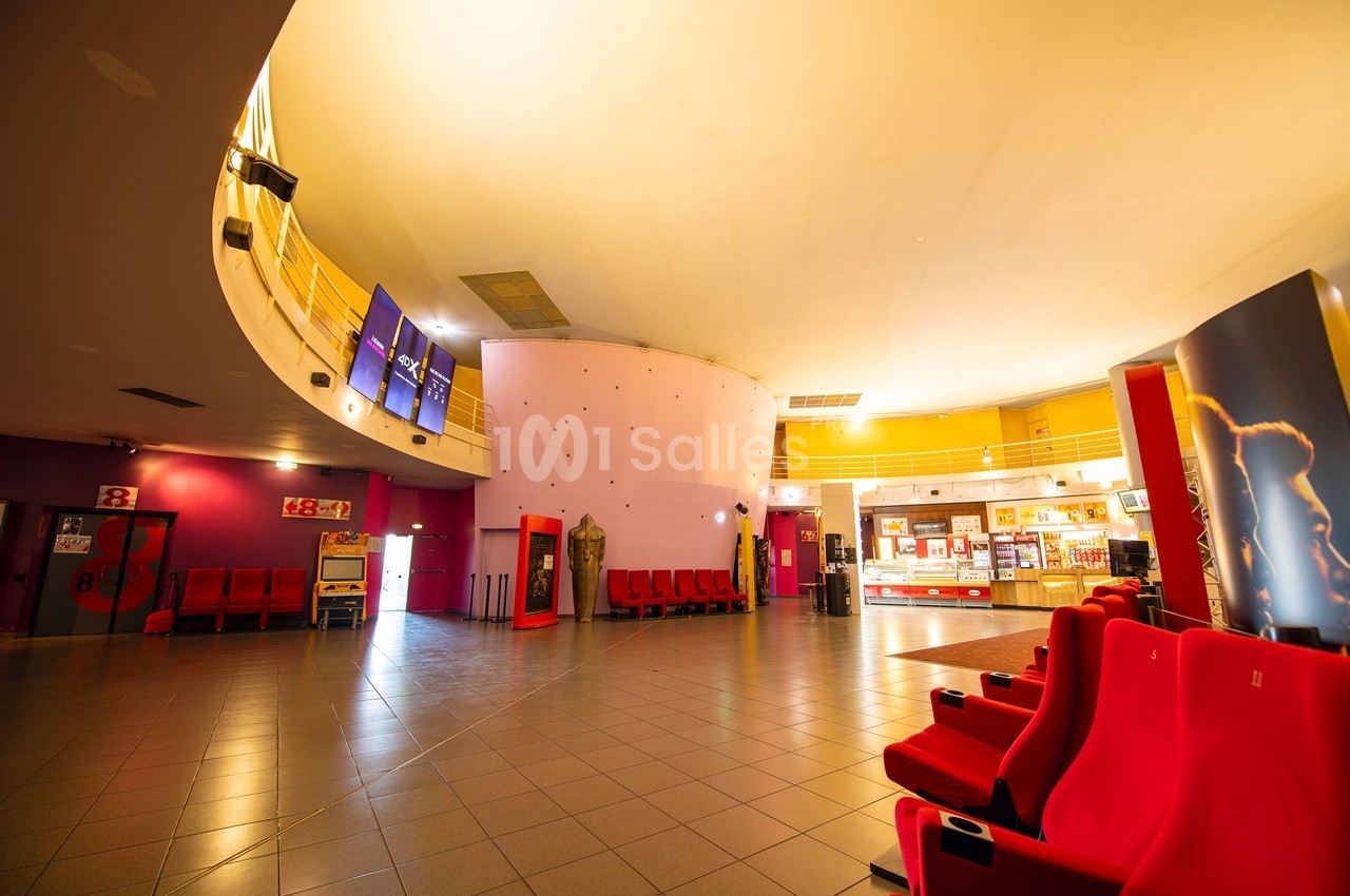 Hall d'entrée d'un cinéma avec fauteuils rouges, comptoir de confiserie éclairé et écrans suspendus.