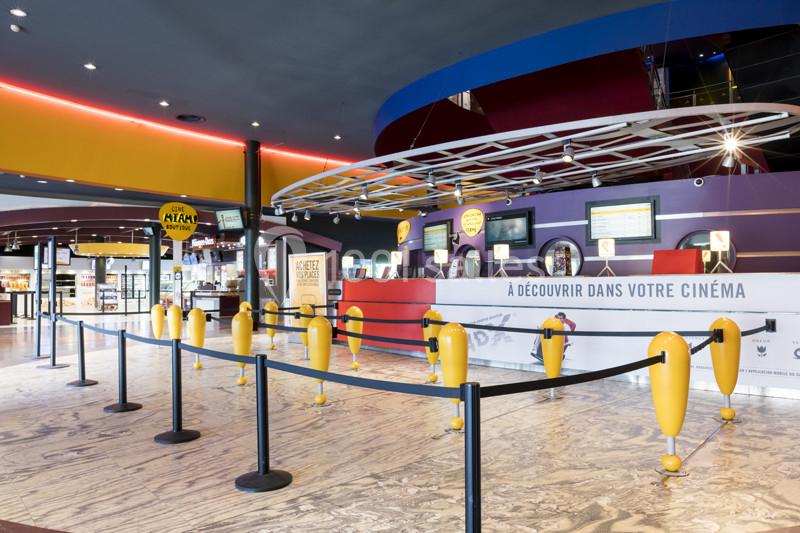 Hall d'entrée d'un cinéma avec guichets, bornes automatiques et files d'attente délimitées par des barrières.