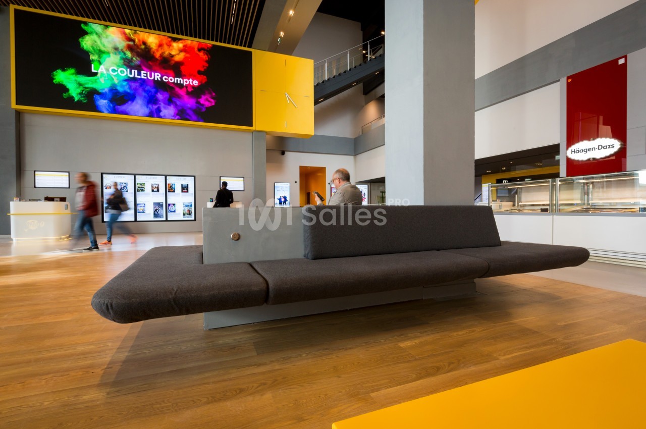Un hall moderne avec un canapé gris, un écran affichant des couleurs vives et des visiteurs en arrière-plan.