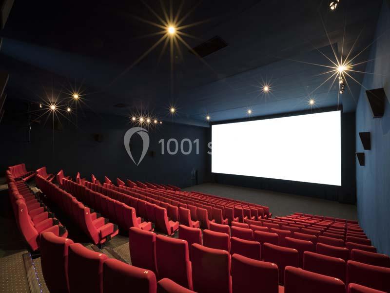 Salle de cinéma vide avec des rangées de sièges rouges et un grand écran blanc allumé.