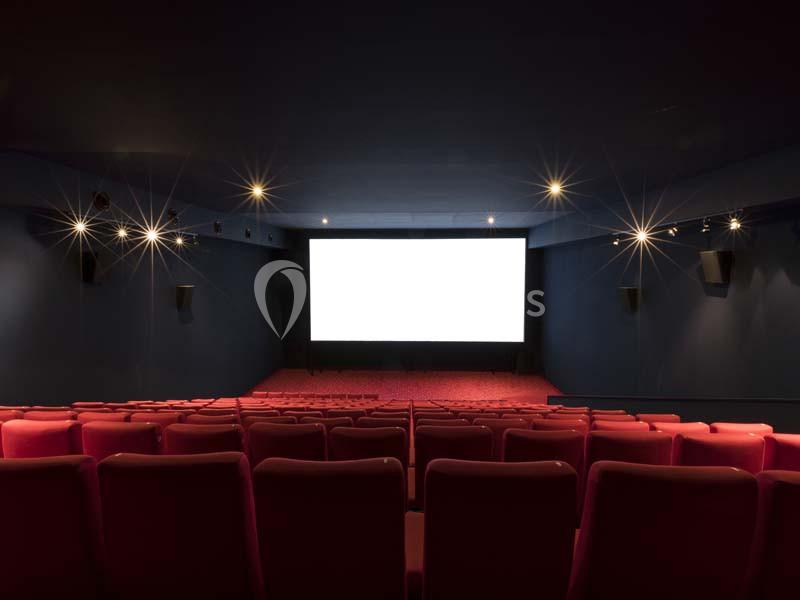 Salle de cinéma vide avec rangées de sièges rouges et un grand écran blanc allumé au fond.