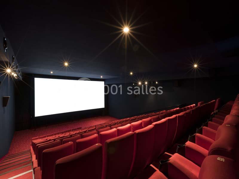 Salle de cinéma vide avec rangées de sièges rouges et grand écran blanc allumé dans une ambiance sombre.